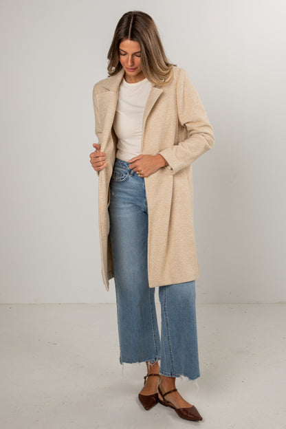 Woman wearing a beige coat, white shirt, and blue jeans on a plain background