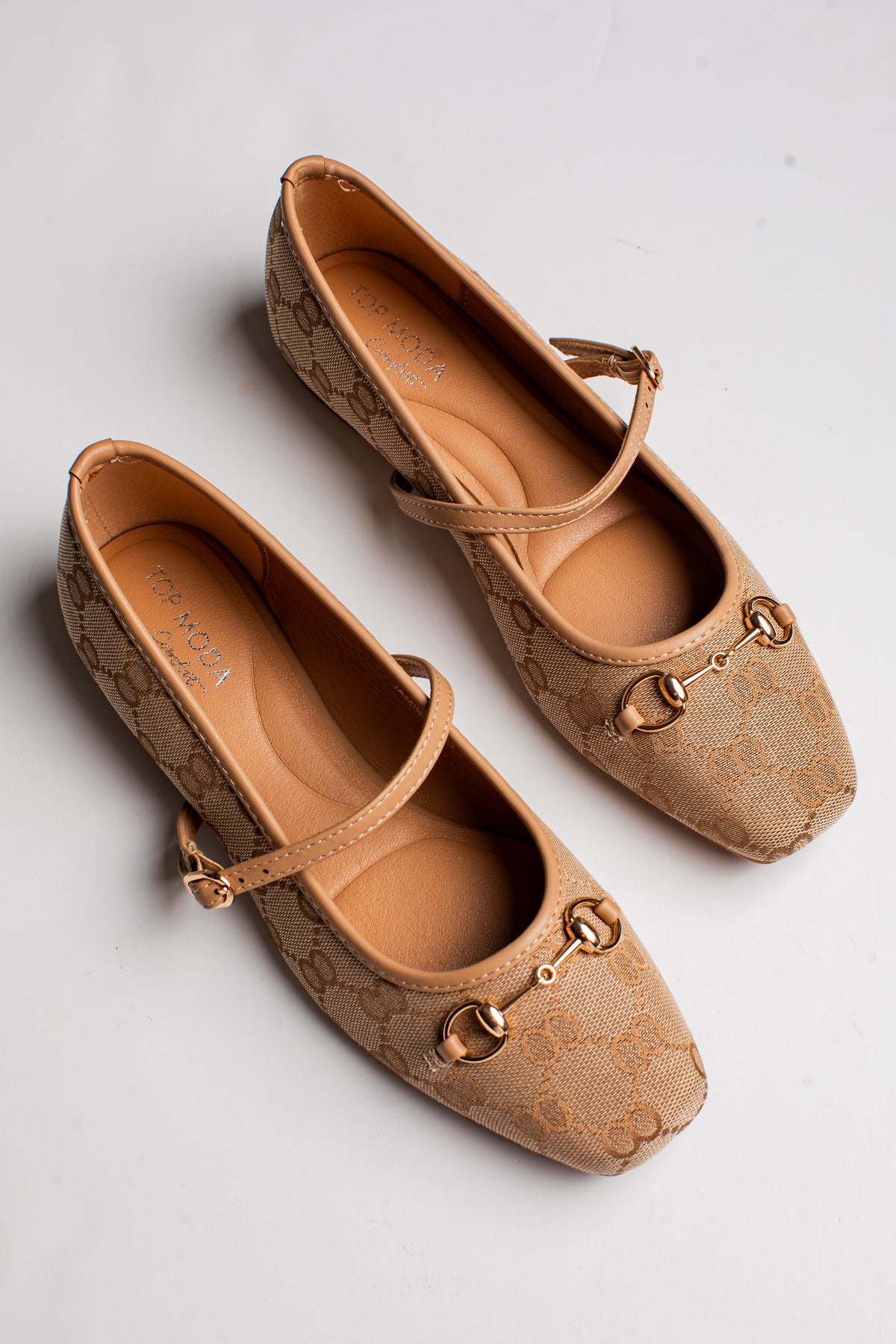 A pair of beige monogram Mary Jane flats with gold-tone horsebit details on the toes, photographed from a top-down angle on a light gray background.