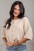A woman with long dark hair wearing a beige short-sleeve button-front top and light blue jeans, standing against a plain light gray wall.