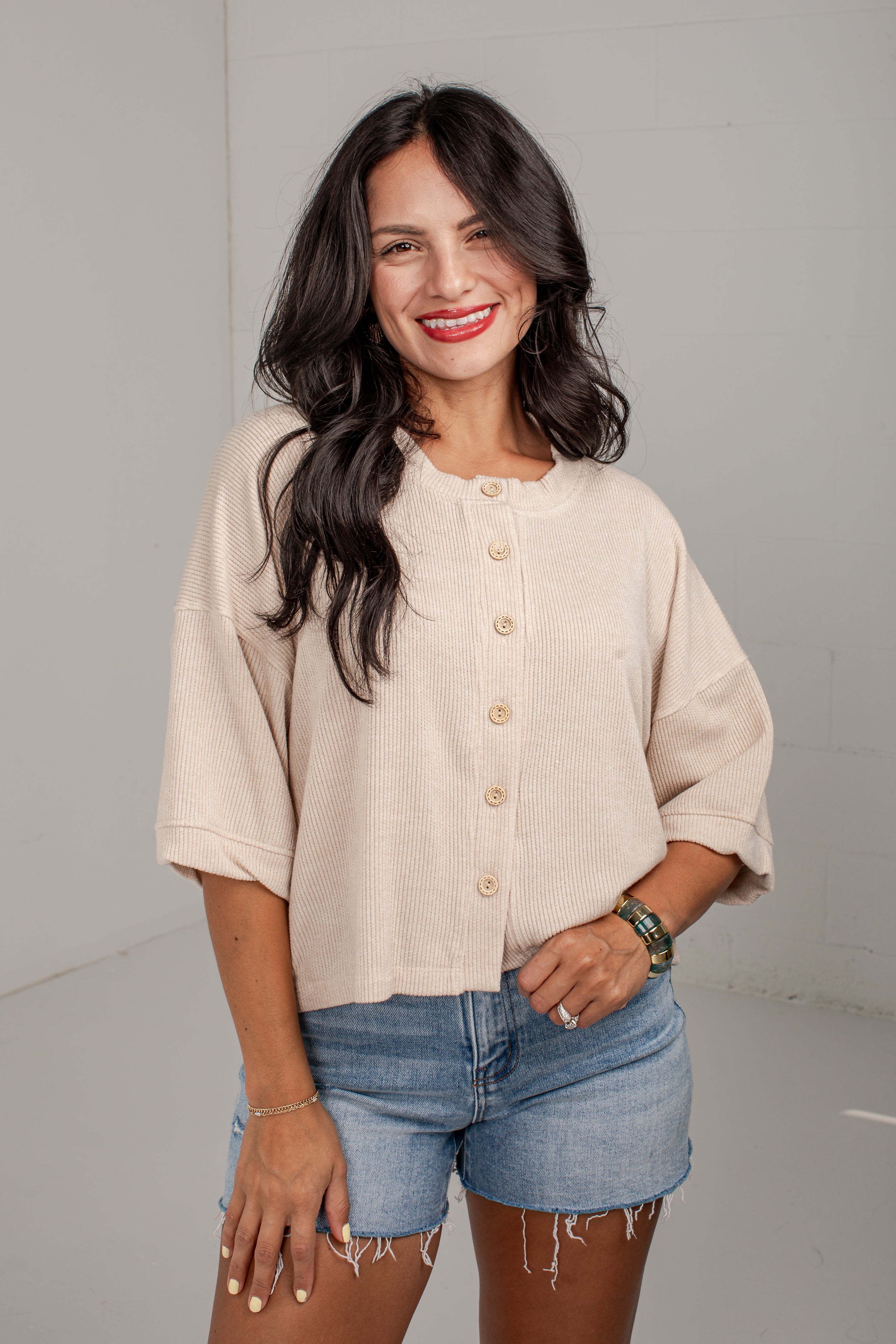 A woman modeling a light beige, short-sleeve button-up top with rolled cuffs and denim shorts against a plain light gray studio background.