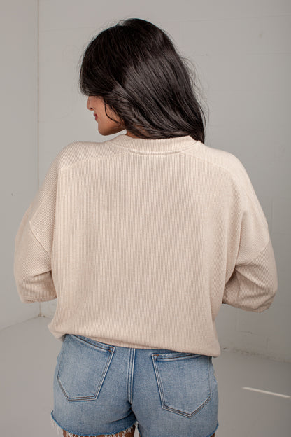A woman shown from behind wearing a light beige ribbed knit long-sleeve top and blue denim jeans in a minimalist studio setting.