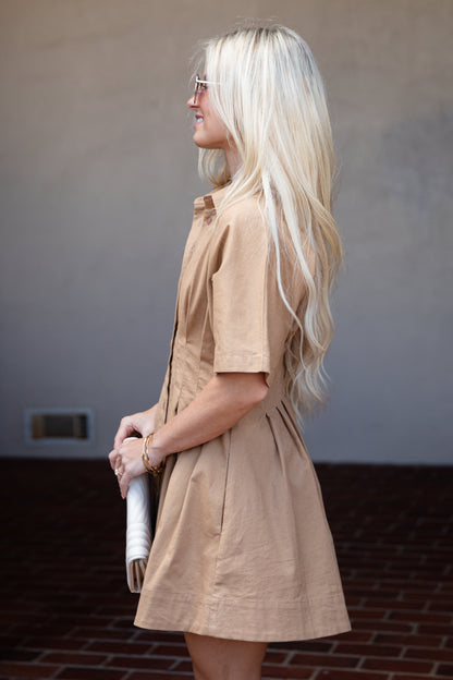 A woman with long blonde hair is shown in profile wearing a short-sleeve tan linen dress, holding a white clutch against a neutral wall and brick floor background.