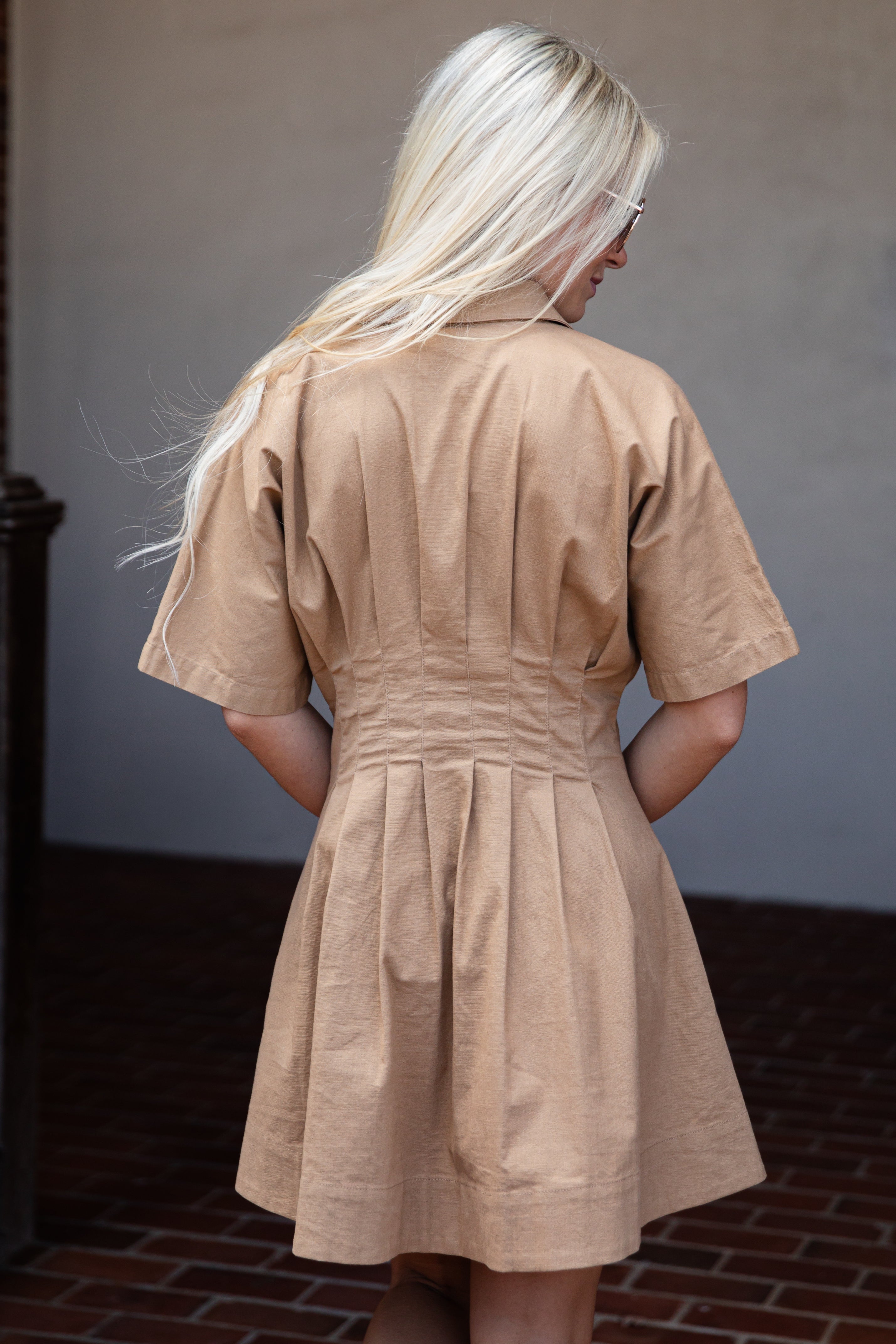 A woman with long blonde hair is shown from behind wearing a tan short-sleeve collared dress with a gathered waist and flared skirt, standing against a neutral wall and brick floor.