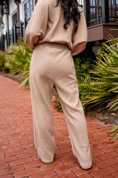 Person wearing a beige outfit standing on a brick path with greenery in the background