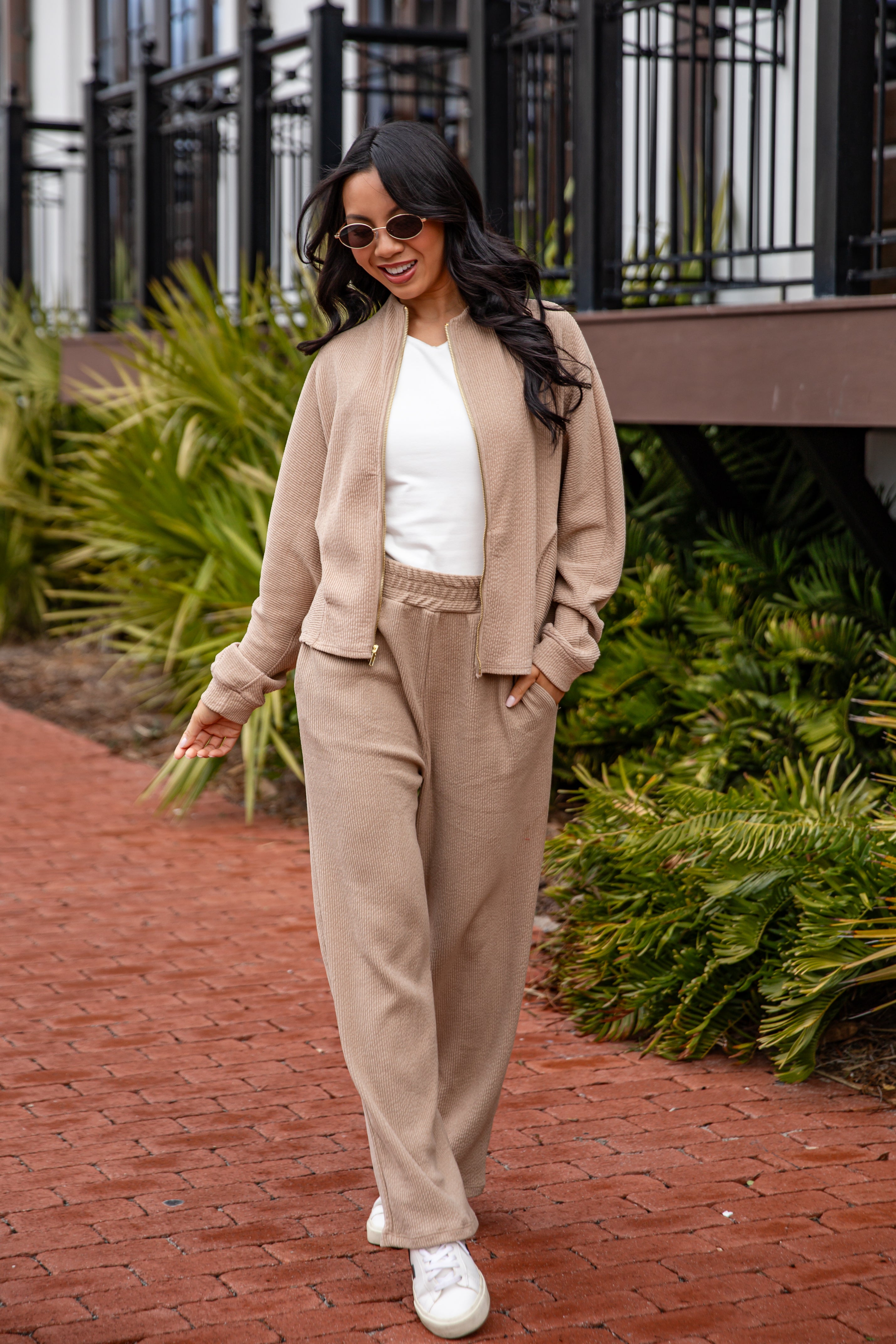 Woman in beige outfit walking on a brick path with greenery in the background
