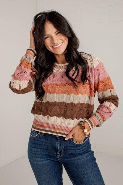 A woman wearing a long-sleeve knit sweater with horizontal stripes in pink, orange, brown, and cream, paired with blue jeans, posing against a white brick wall.