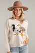 A woman wearing a beige knit sweater with a large embroidered duck family motif, paired with light blue jeans and a brown wide-brim hat, standing against a light gray wall.