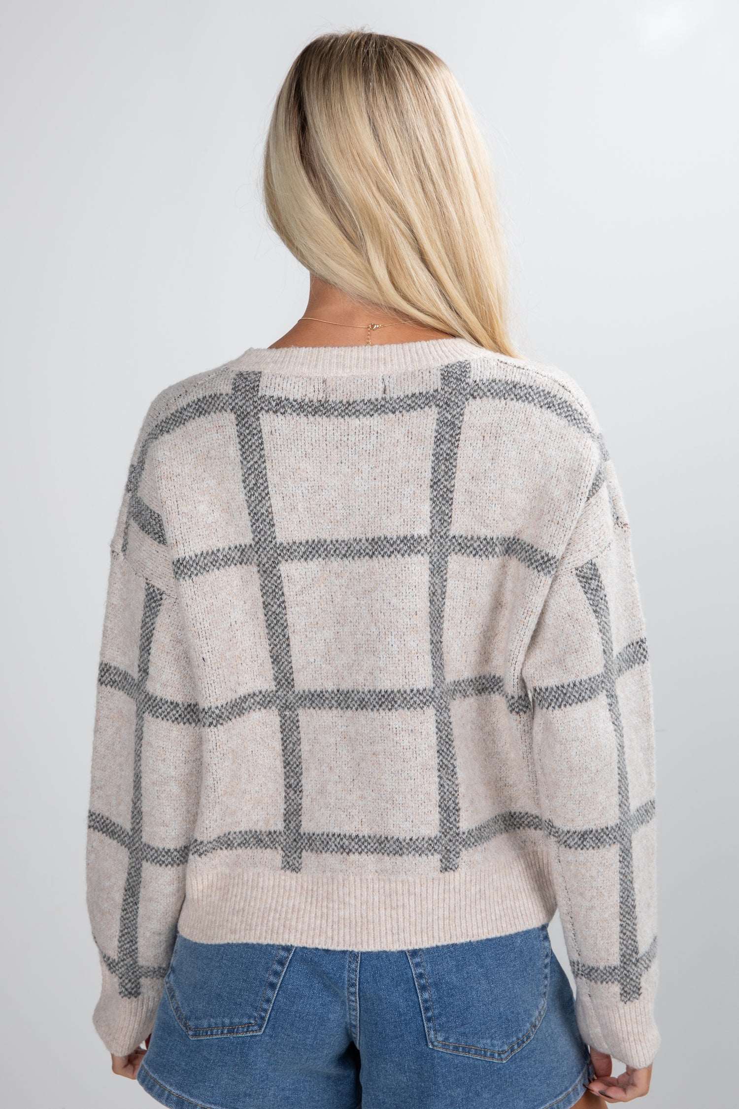 Person wearing a beige sweater with black grid pattern and blue jeans on a white background