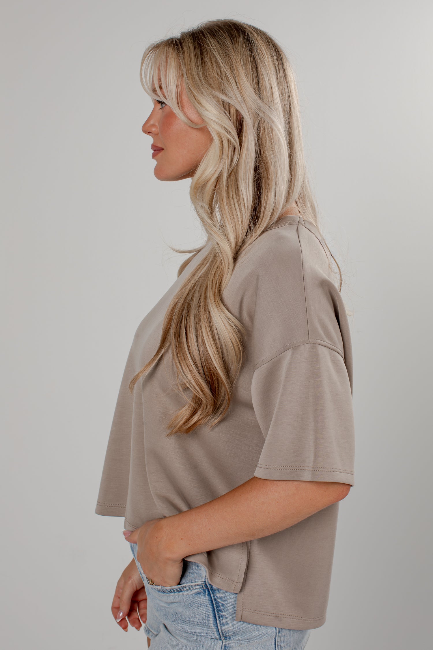 A woman with long blonde hair is shown in profile wearing a taupe short-sleeve crop top and light blue jeans against a plain light gray studio background.