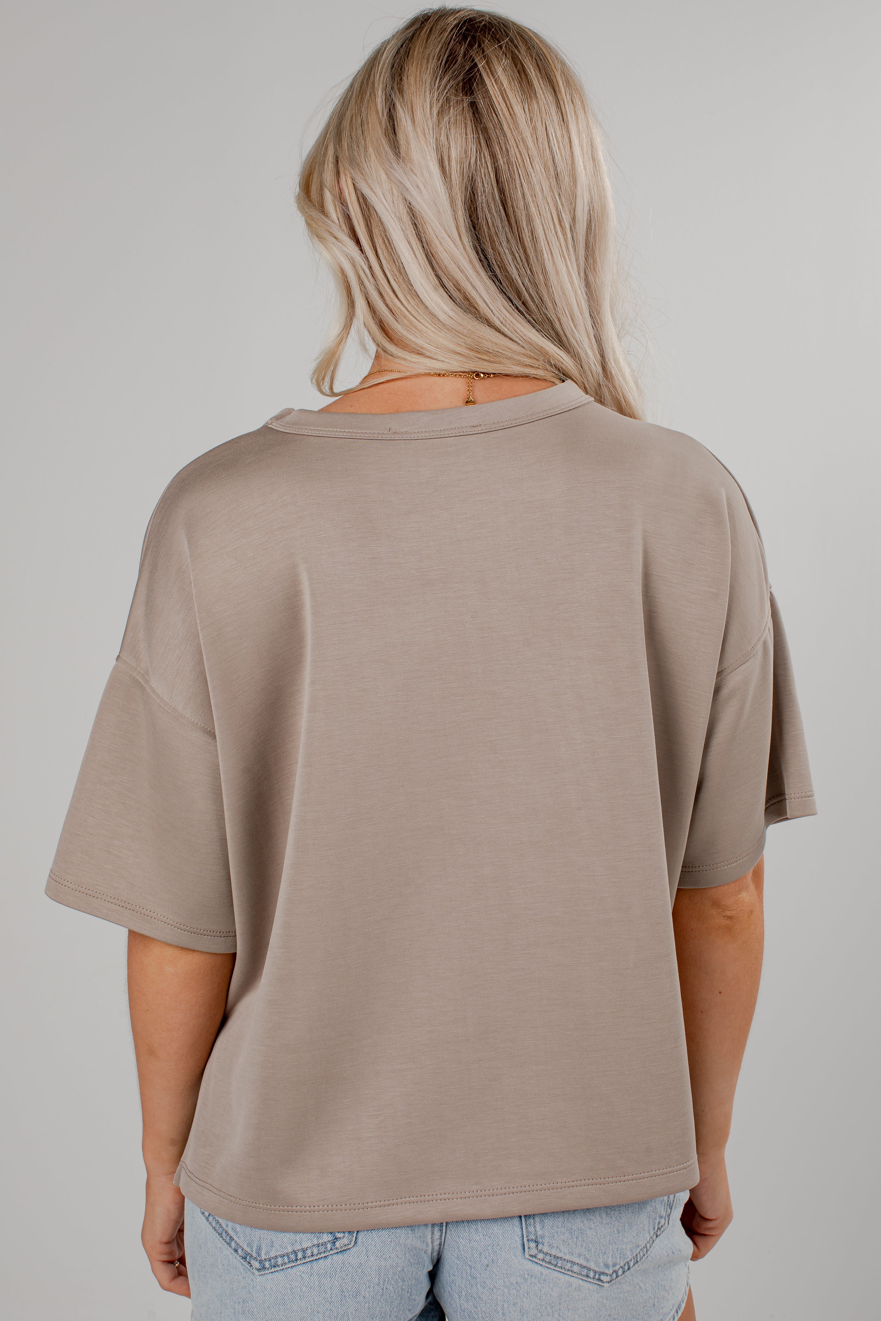 A woman viewed from the back wearing a loose-fit taupe short-sleeve t-shirt and light blue jeans against a solid light gray background.