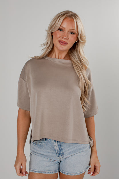 A blonde woman wearing a taupe short-sleeve crewneck t-shirt and light-wash denim shorts, standing against a neutral gray studio background.