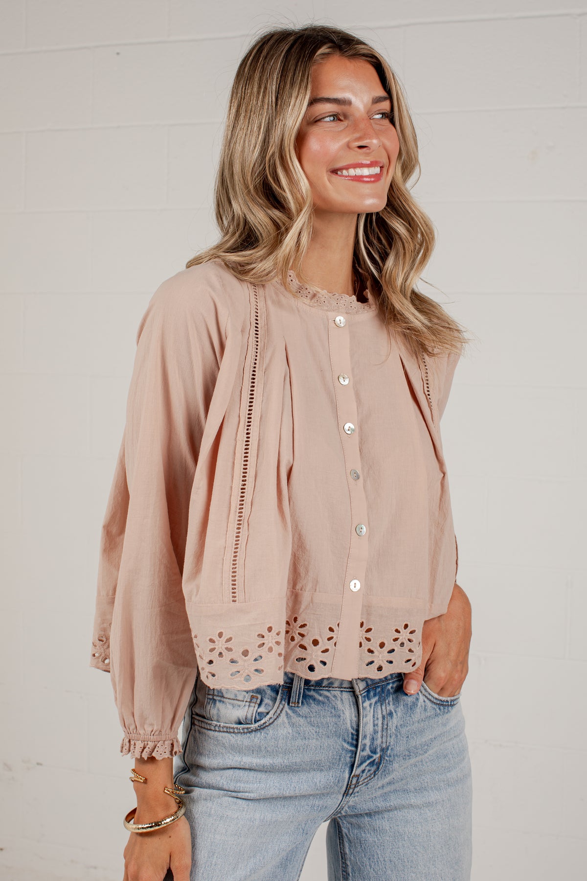 A woman wearing a light dusty pink eyelet blouse with button-front closure and gathered sleeves, paired with light-wash blue jeans, standing against a white paneled wall.
