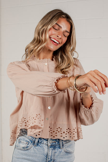 A woman wearing a light beige long-sleeve blouse with eyelet lace detailing at the hem and ruffled cuffs, paired with light blue jeans, posing in front of a white brick wall.