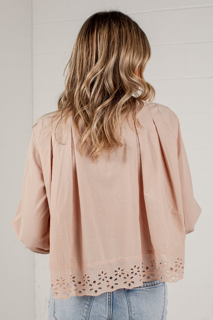 A woman viewed from behind wearing a dusty pink blouse with pleated shoulders and eyelet lace trim at the hem, paired with light blue jeans against a white brick wall.