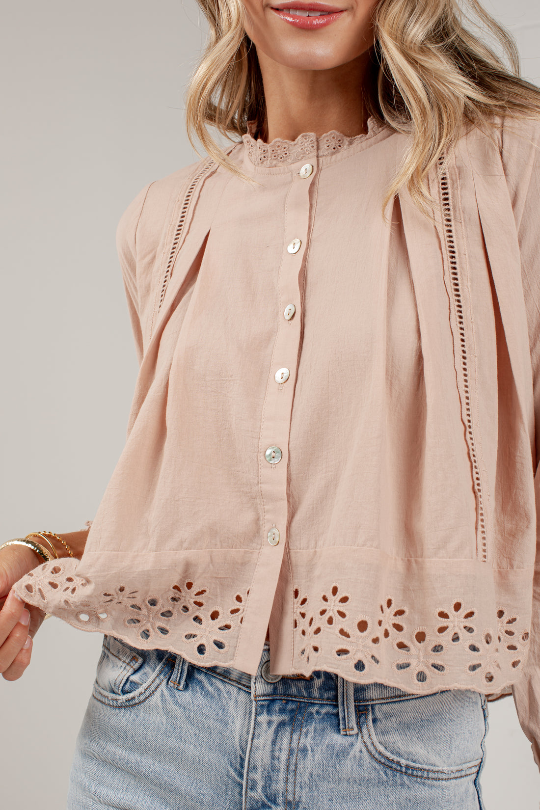 A woman wearing a light blush pink blouse with eyelet lace trim at the collar and hem, button-front closure, and pleated side details, paired with light-wash blue jeans against a plain light gray studio background.