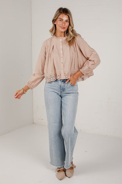 A woman modeling a light blush pink long-sleeve blouse with eyelet lace trim and button details, paired with light-wash wide-leg jeans and beige pointed flats, standing against a white brick wall.
