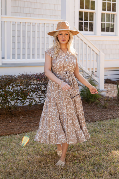 Woman in a patterned dress and hat standing in front of a house.