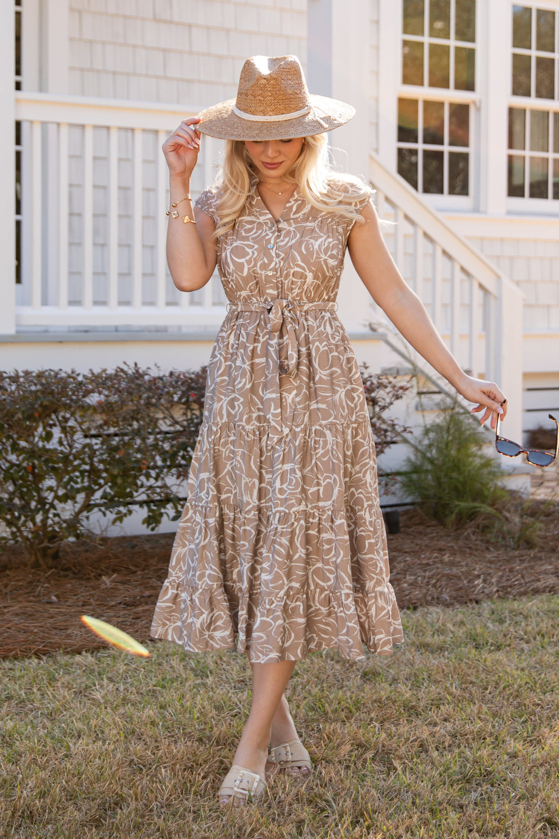 Woman in a floral dress and hat standing in front of a white house.