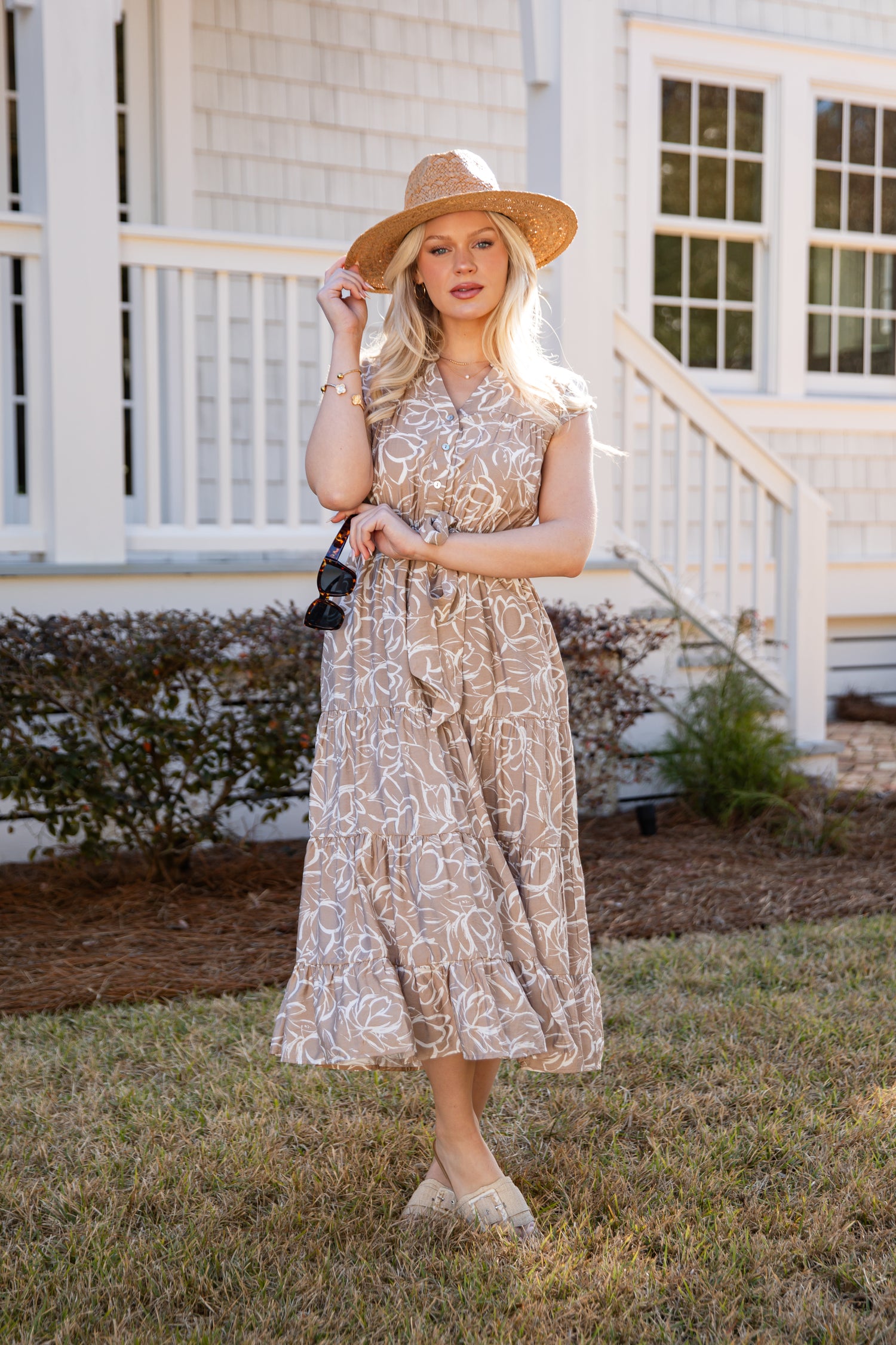 Woman in a patterned dress and hat standing in front of a house.