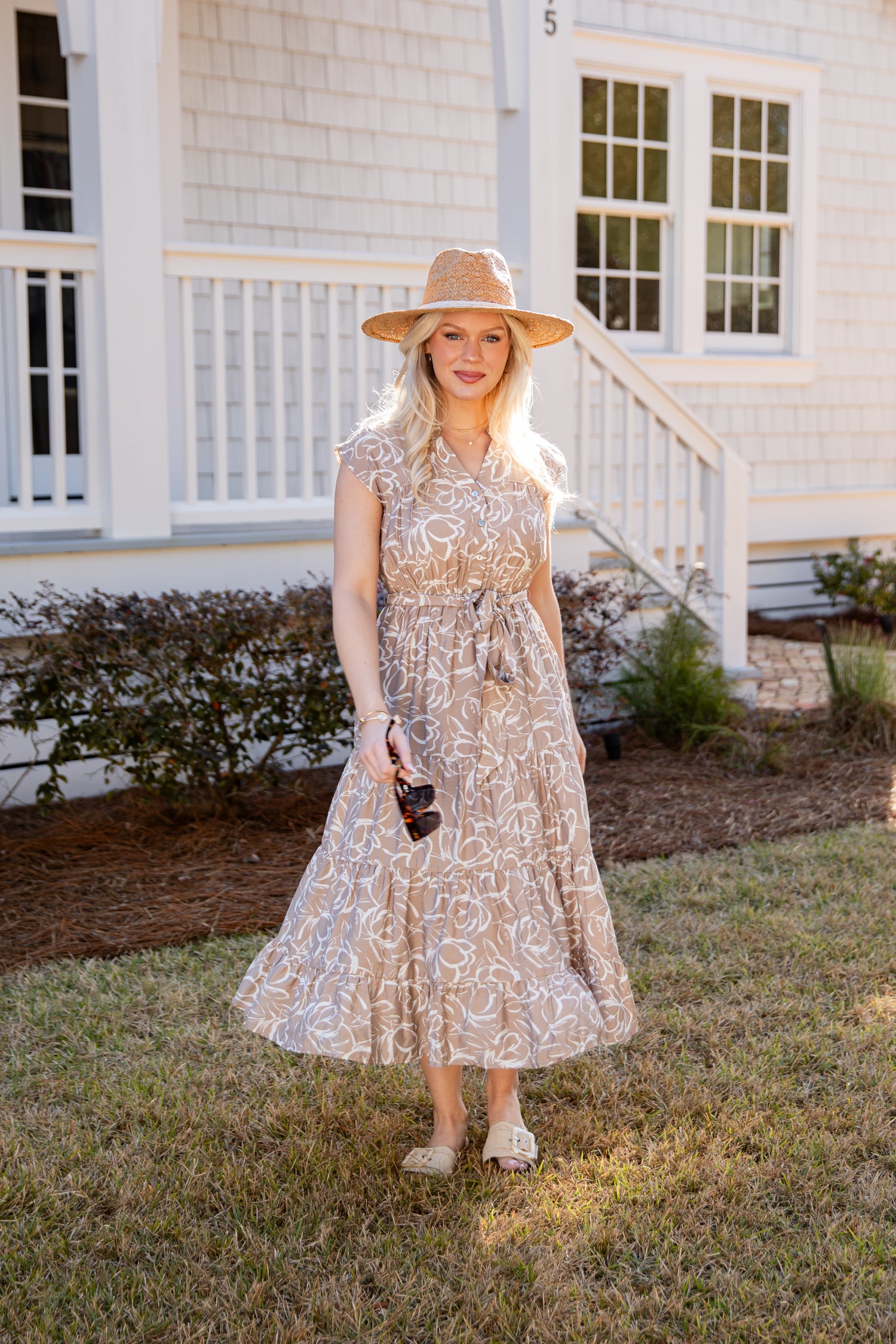 Woman in a floral dress and hat standing in front of a white house.