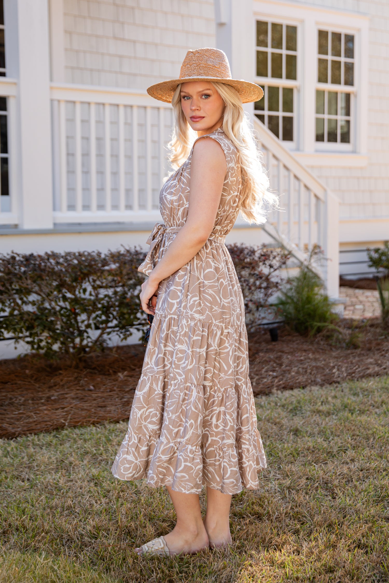 Woman in a patterned dress and hat standing in front of a house.