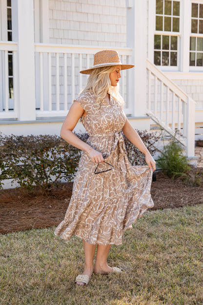 Woman in a patterned dress and straw hat standing in front of a white house.