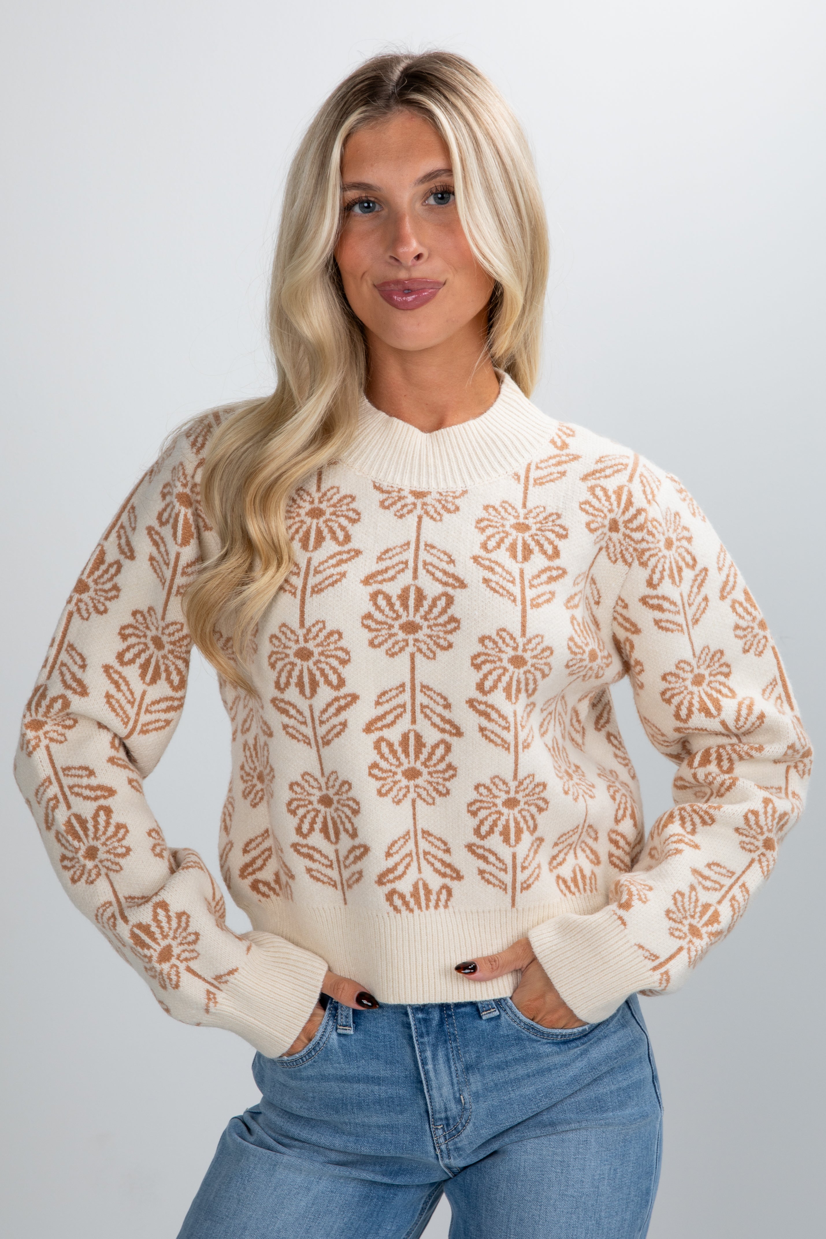 Woman wearing a cream sweater with brown floral patterns and blue jeans on a plain background
