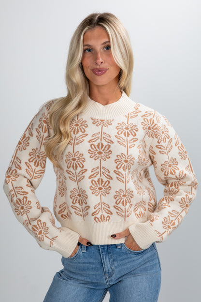 Woman wearing a cream sweater with brown floral patterns and blue jeans on a plain background