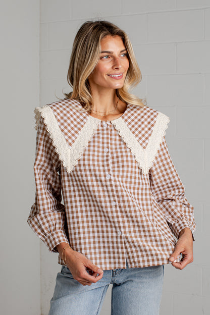 A woman wearing a brown and white gingham long-sleeve blouse with a large cream lace collar, paired with light blue jeans, standing against a light gray brick wall.