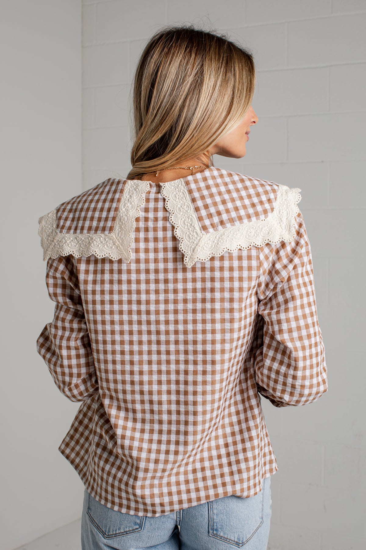 A woman viewed from the back wearing a brown and beige gingham long-sleeve blouse with a large cream lace collar, paired with light blue jeans, set against a white brick wall.