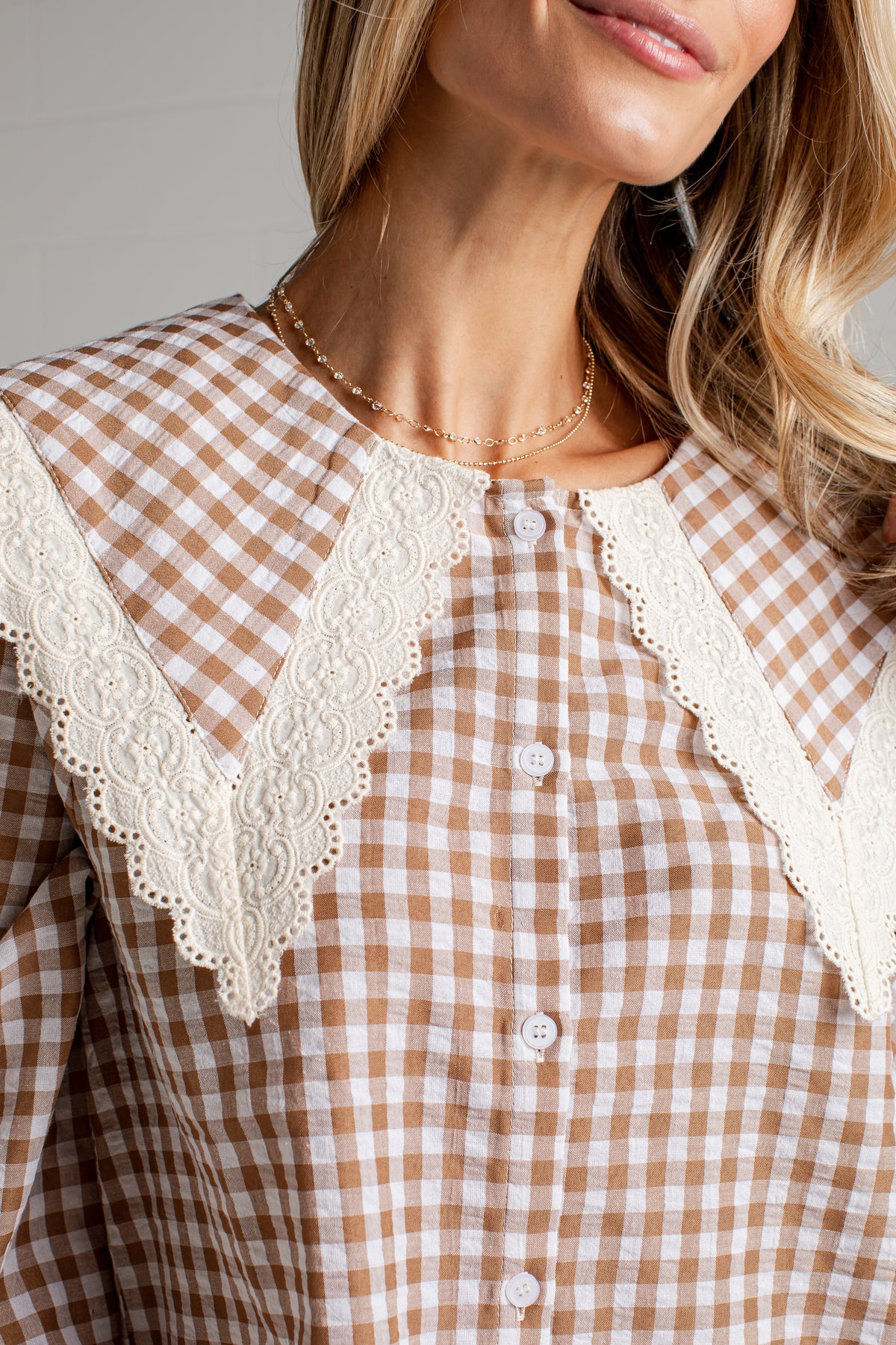 A close-up of a woman wearing a brown and beige gingham button-up shirt with a large cream lace collar.