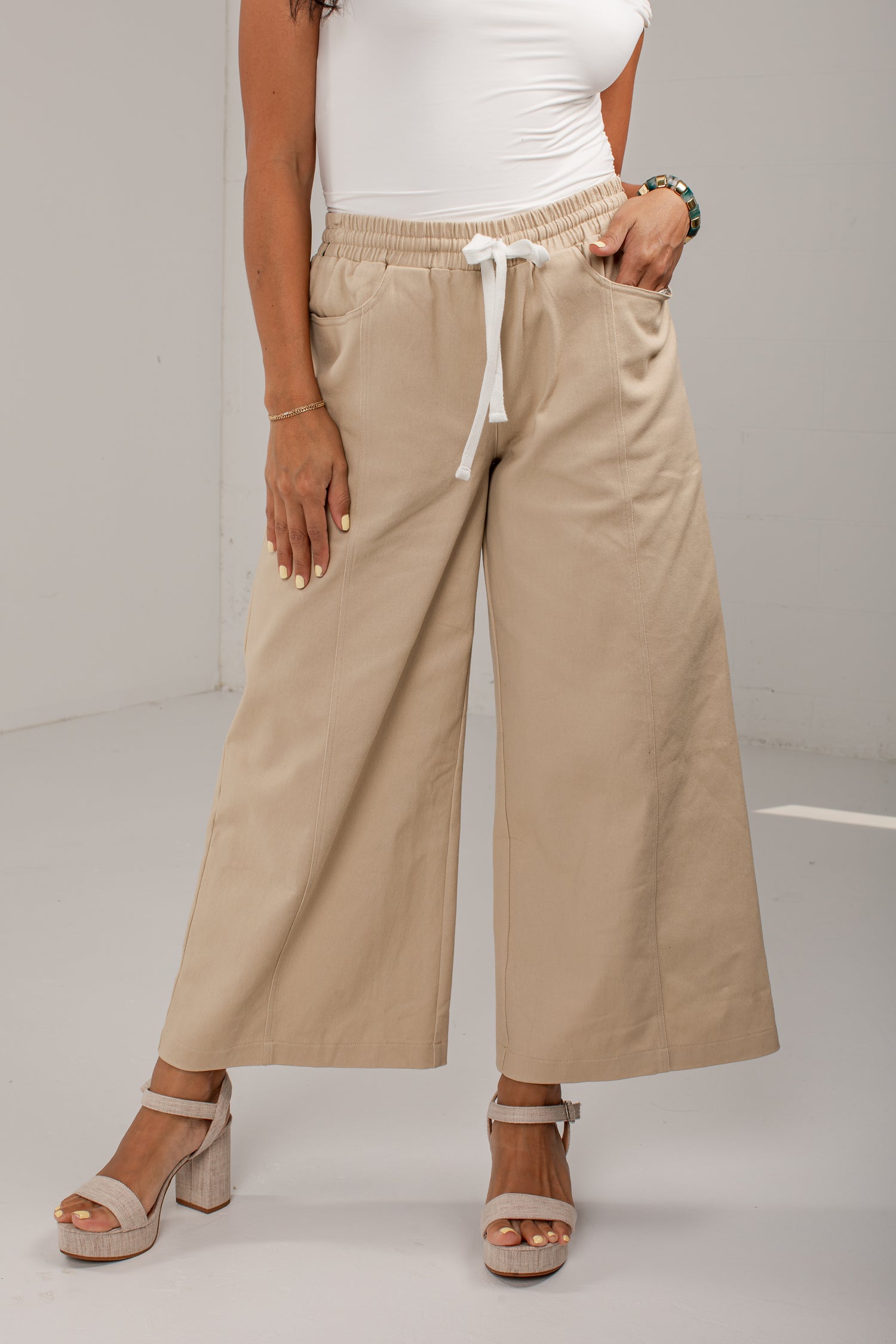 A model wearing beige wide-leg cropped pants with an elastic waist and white drawstring, paired with a white top and beige platform sandals.