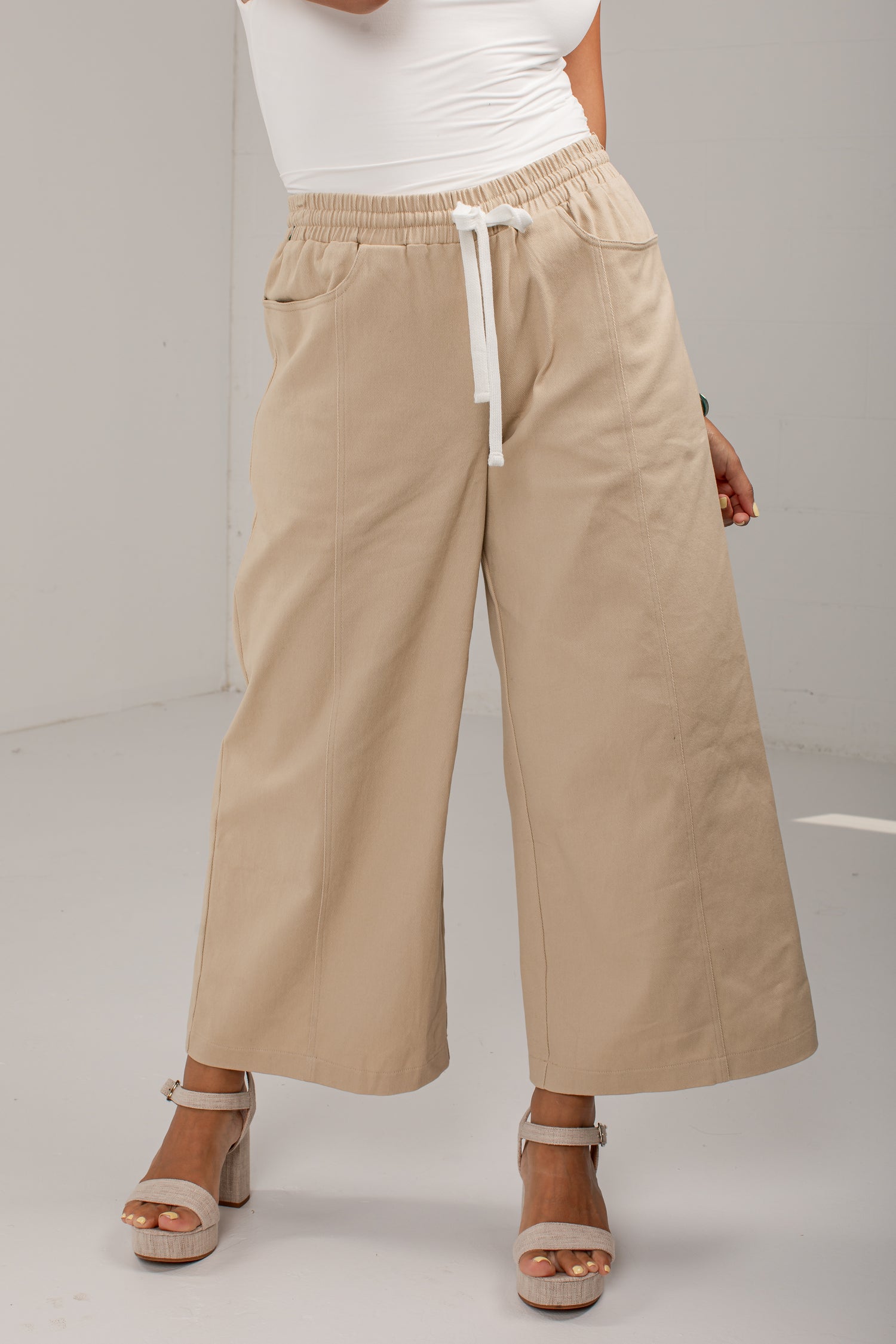 A person wearing beige wide-leg cropped pants with an elastic waistband and white drawstring, paired with a white top and beige heeled sandals, photographed in a studio setting.