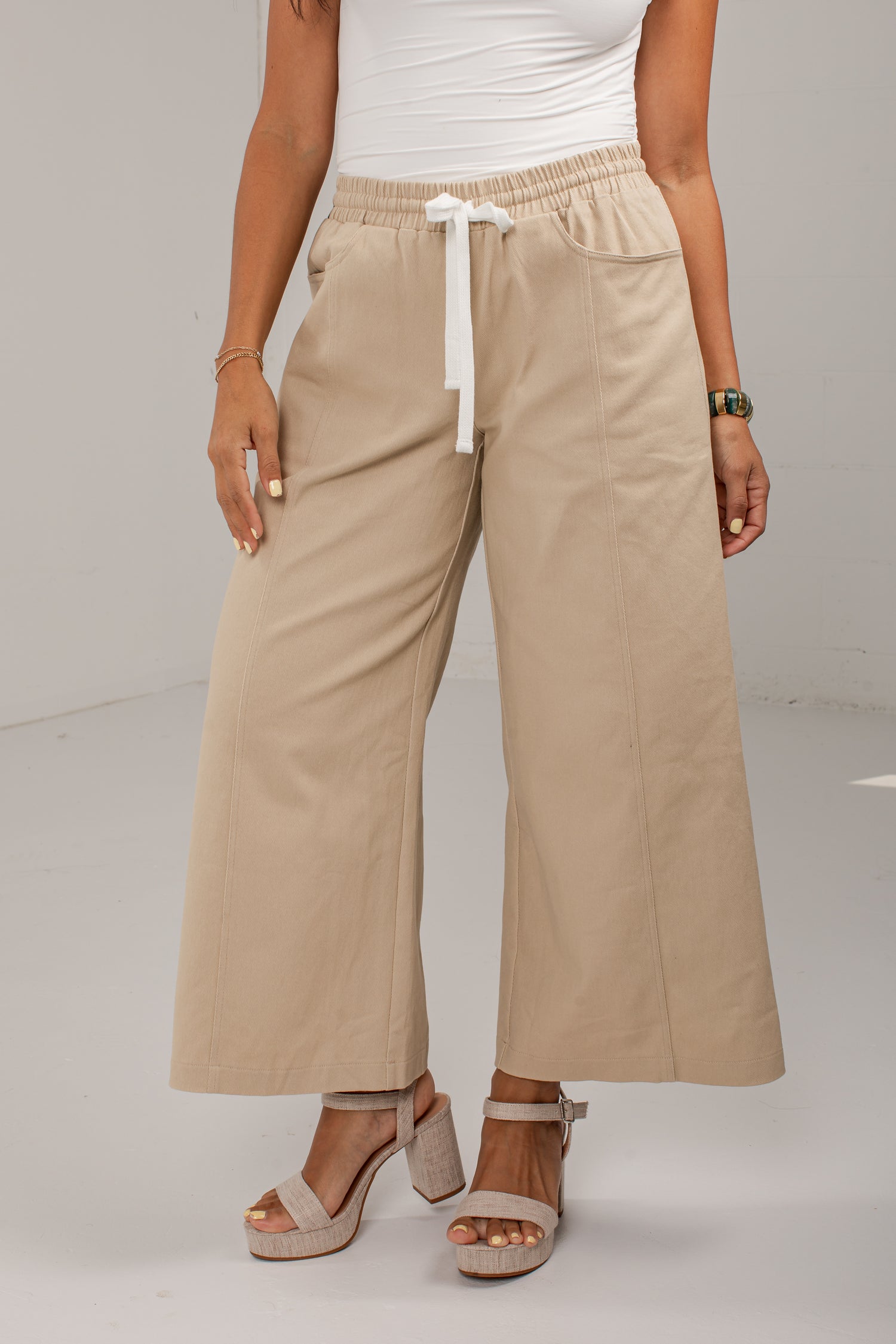 A model wearing beige wide-leg cropped pants with an elastic waistband and white drawstring, paired with a white top and beige platform sandals.
