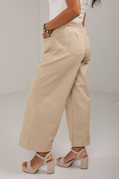A person wearing beige wide-leg cropped pants with an elastic waistband, shown from the back in a studio setting. The model is also wearing a white top and beige block-heel sandals.
