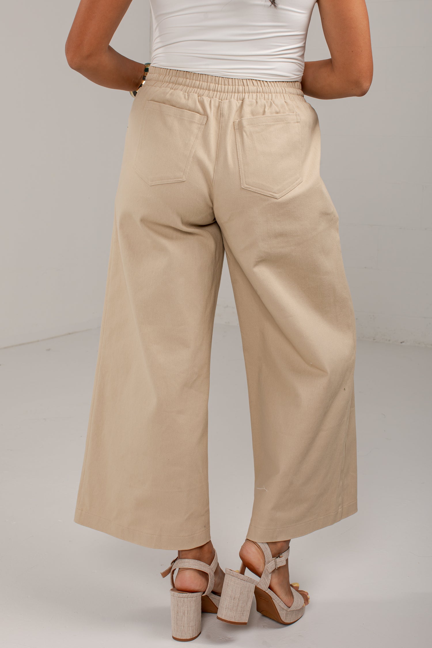 A person shown from the back wearing wide-leg cropped pants in a light beige or sand color, paired with a white top and beige heeled sandals, against a plain light gray studio background.