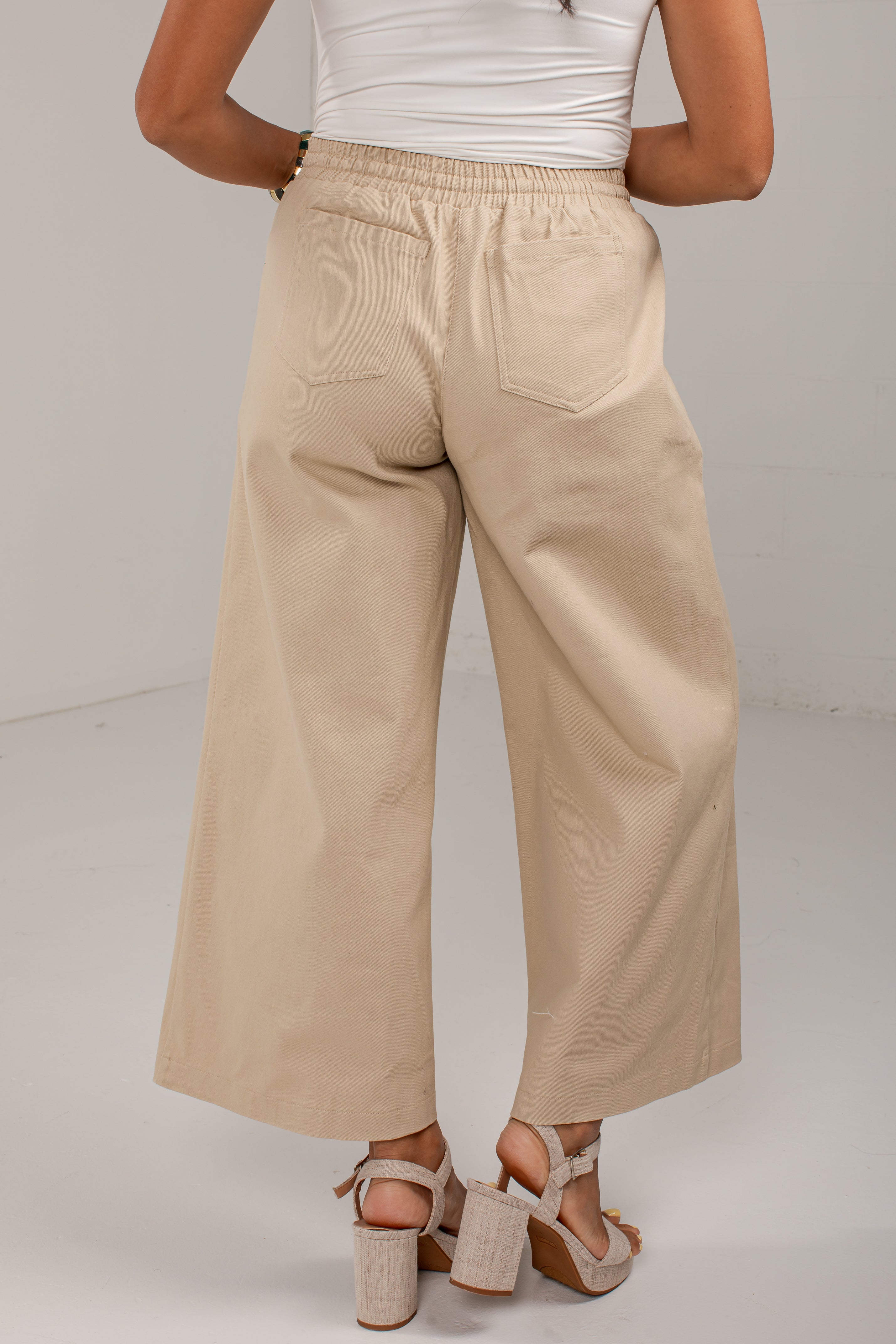 A person shown from the back wearing wide-leg cropped pants in a light beige or sand color, paired with a white top and beige heeled sandals, against a plain light gray studio background.
