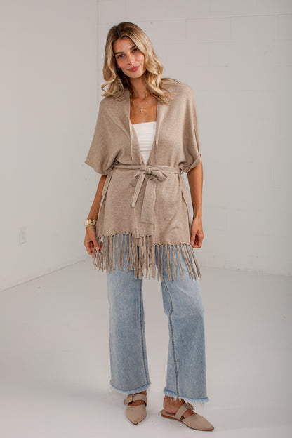 A woman models a taupe-beige short-sleeve fringe cardigan with a tie waist, worn over a white tank top and light-wash wide-leg jeans.