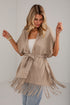 A woman wearing a beige wrap poncho with fringe trim and a tie belt, styled over a white top and light blue jeans, set against a light gray studio background.