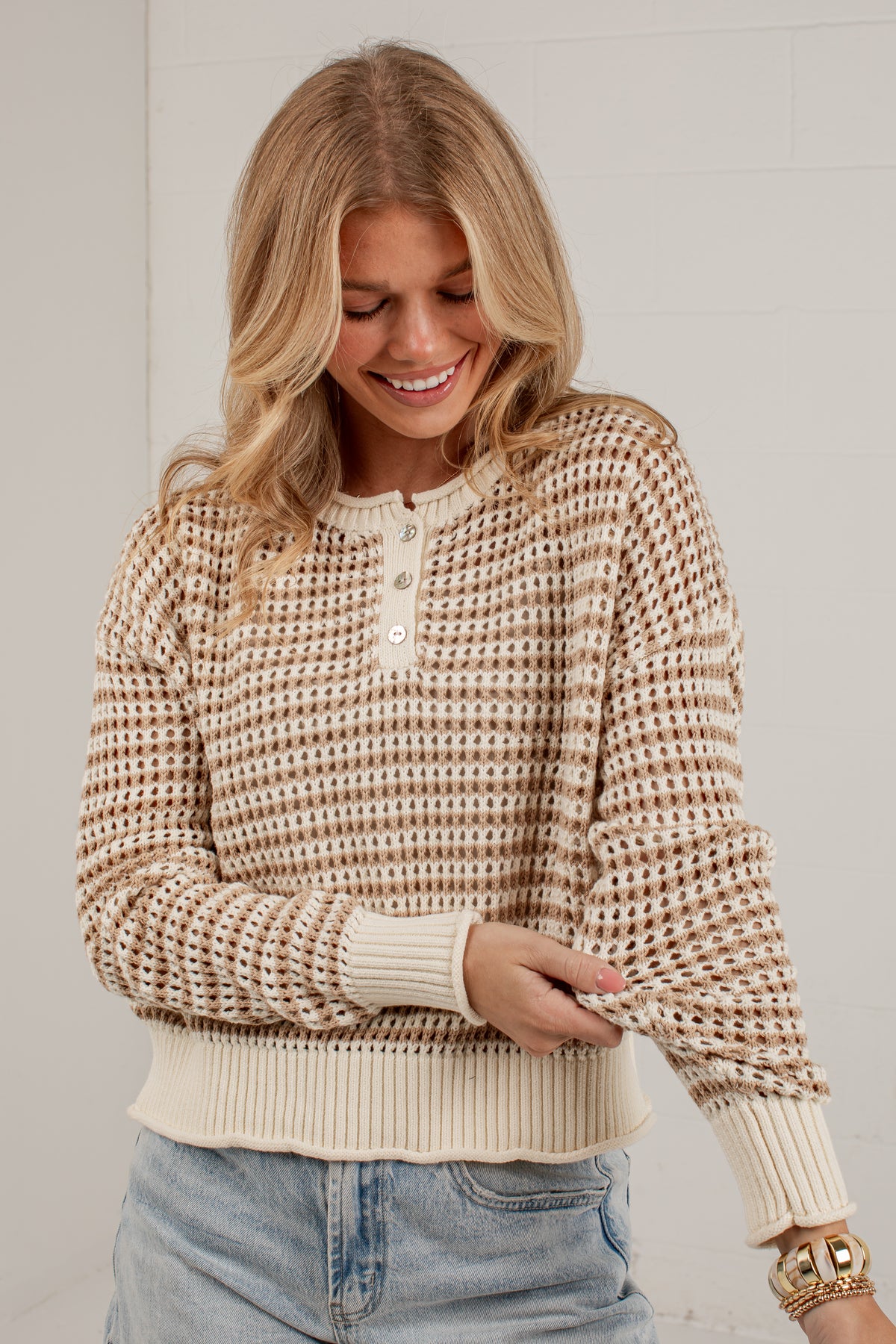 A woman wearing a beige and brown textured knit sweater with a buttoned henley collar, paired with light blue jeans, standing against a white brick wall.
