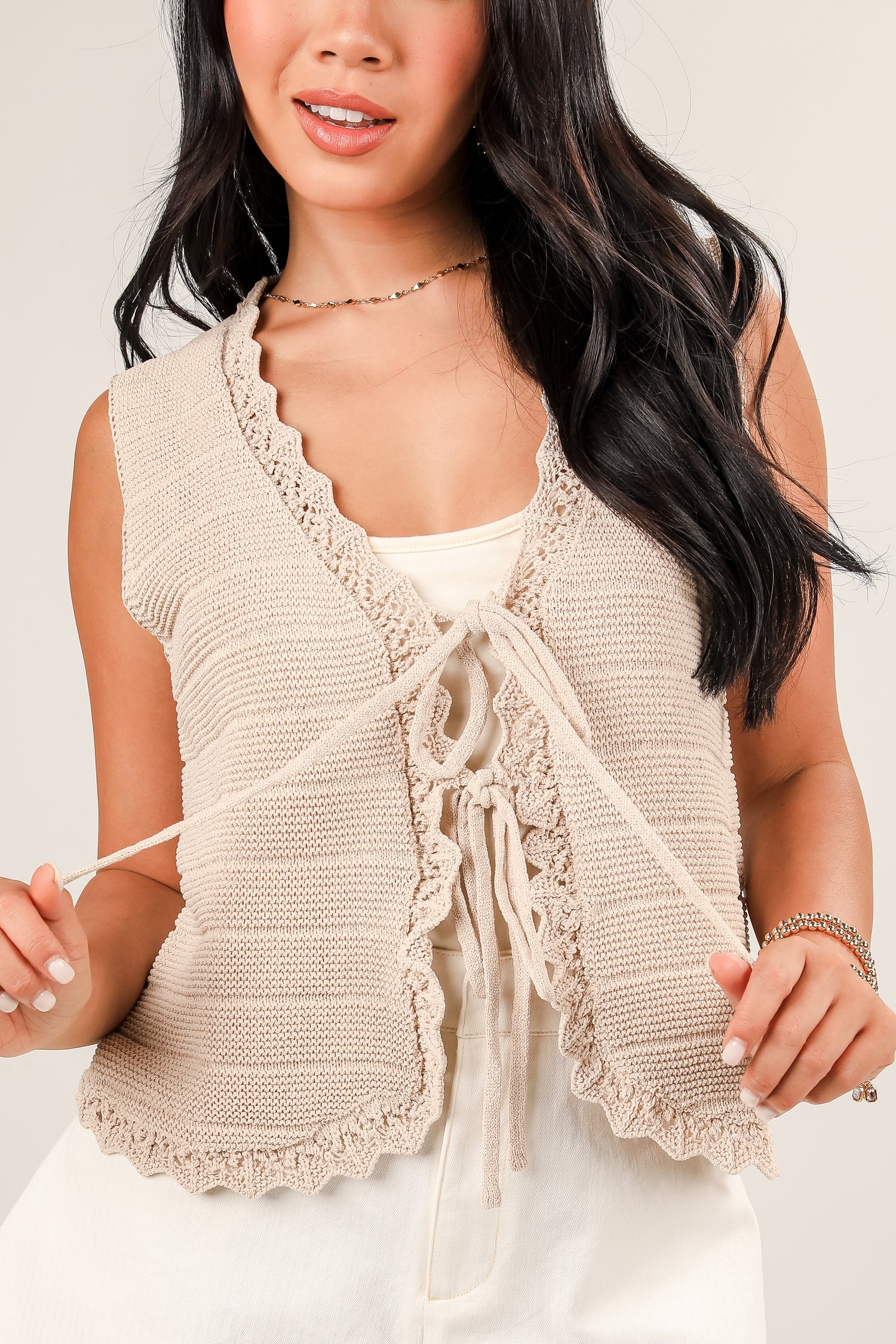 Woman wearing a beige knitted vest over a white top against a plain background