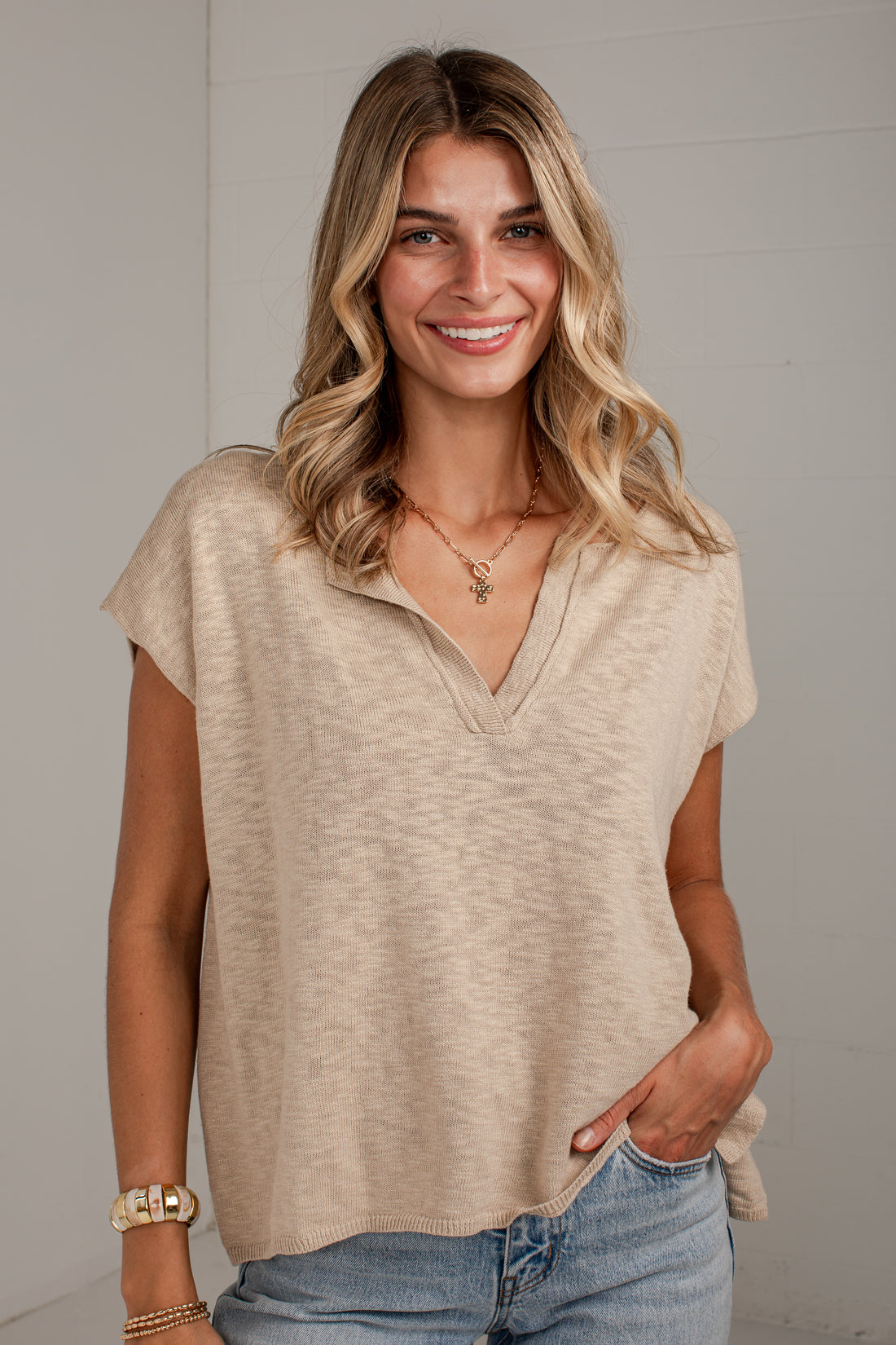 A woman with blonde hair wearing a beige short-sleeve waffle-knit top with a split V-neck, paired with light blue jeans, standing against a light gray paneled wall.