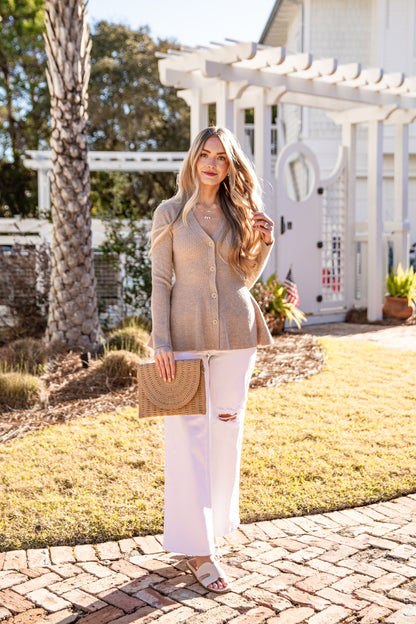 Woman in beige cardigan and white pants holding a straw bag outdoors.