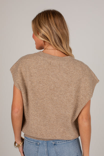 A woman viewed from the back wearing a light brown knit sweater with wide cap sleeves and light blue jeans, set against a plain light gray studio background.