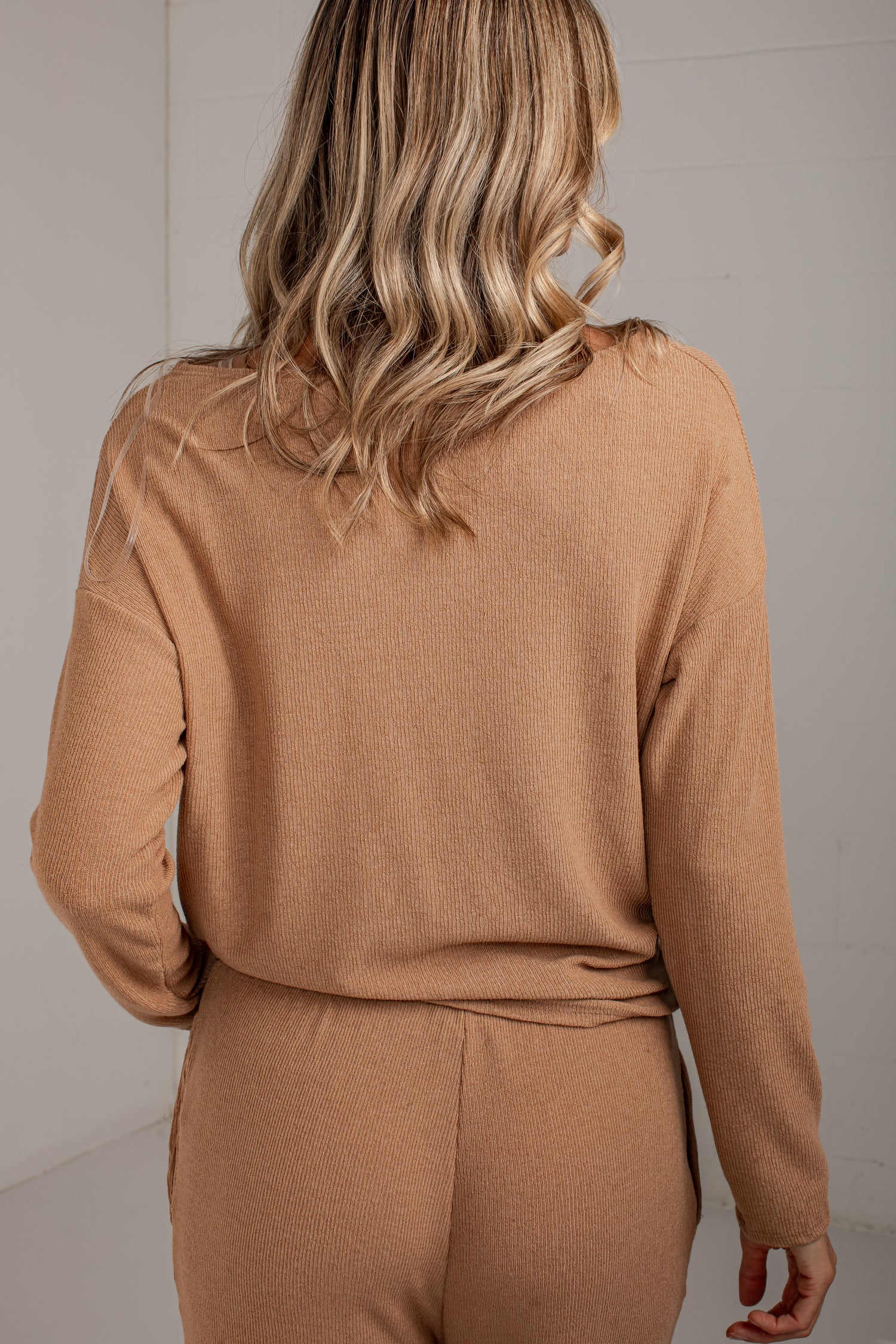 A woman shown from behind wearing a camel-colored knit long-sleeve top and matching pants against a light neutral wall.