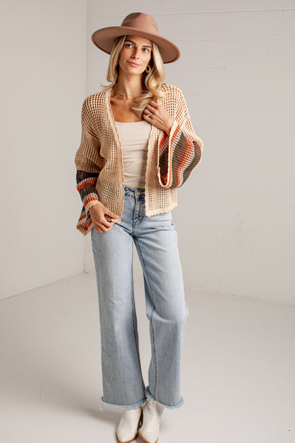 A woman stands in a bright studio wearing a beige wide-brim hat, a cream tank top, a beige crochet cardigan with multicolor striped cuffs, light-wash wide-leg jeans, and white ankle boots.