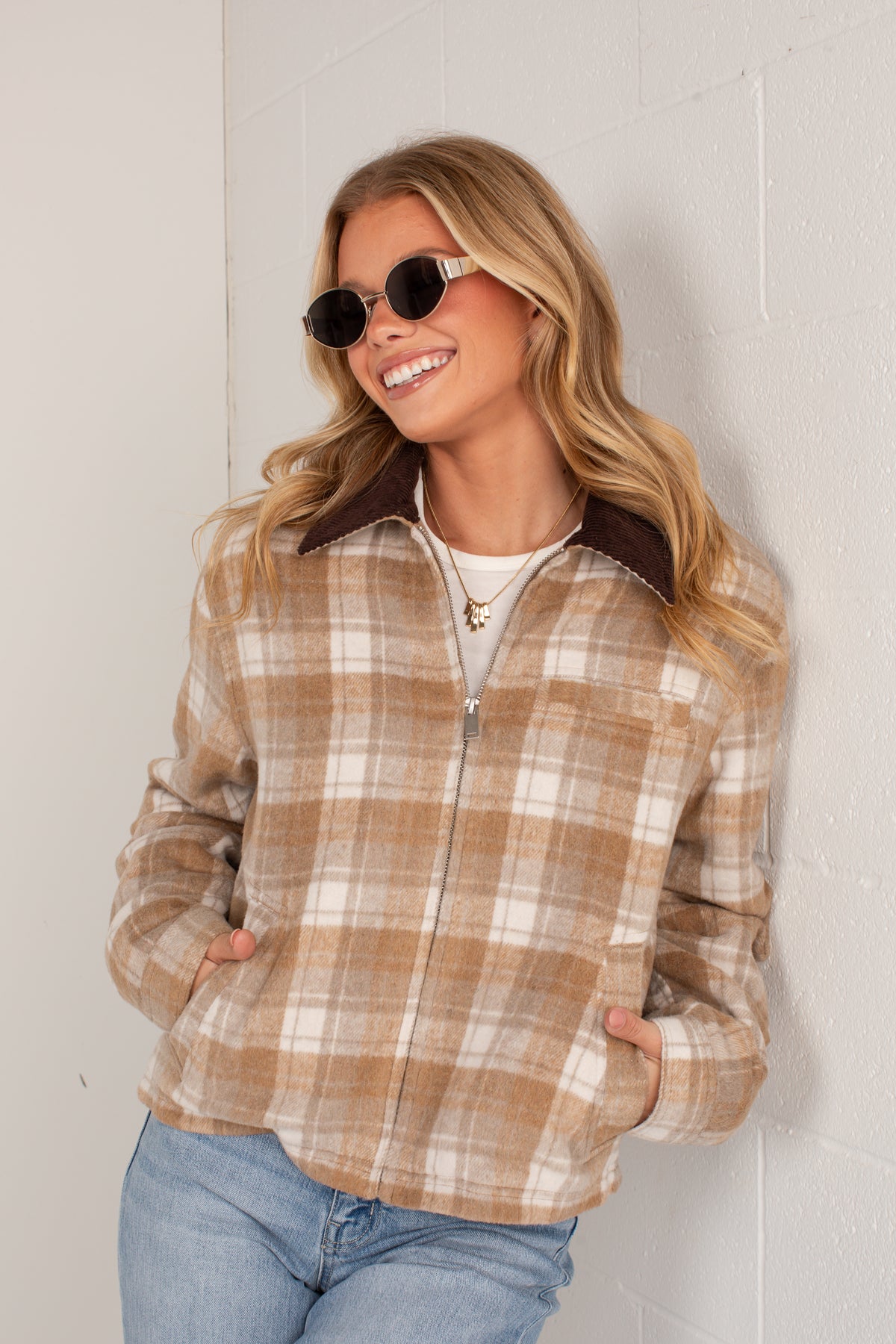 A woman wearing a tan and cream plaid zip-up jacket with a dark brown collar, paired with blue jeans and sunglasses, posing against a white brick wall.