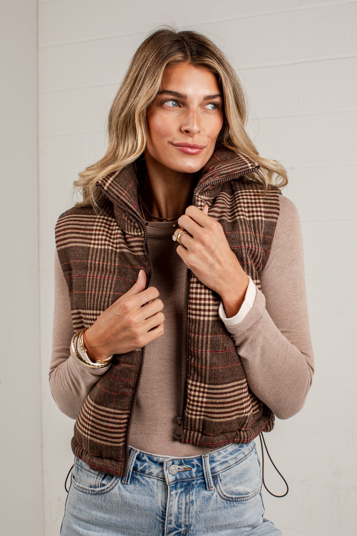 A woman wearing a brown and beige plaid puffer vest over a beige long-sleeve top and blue jeans, standing against a white paneled wall.
