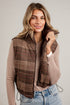 A woman wearing a brown and beige plaid zip-up vest over a beige long-sleeve top and blue jeans, standing against a white paneled wall.