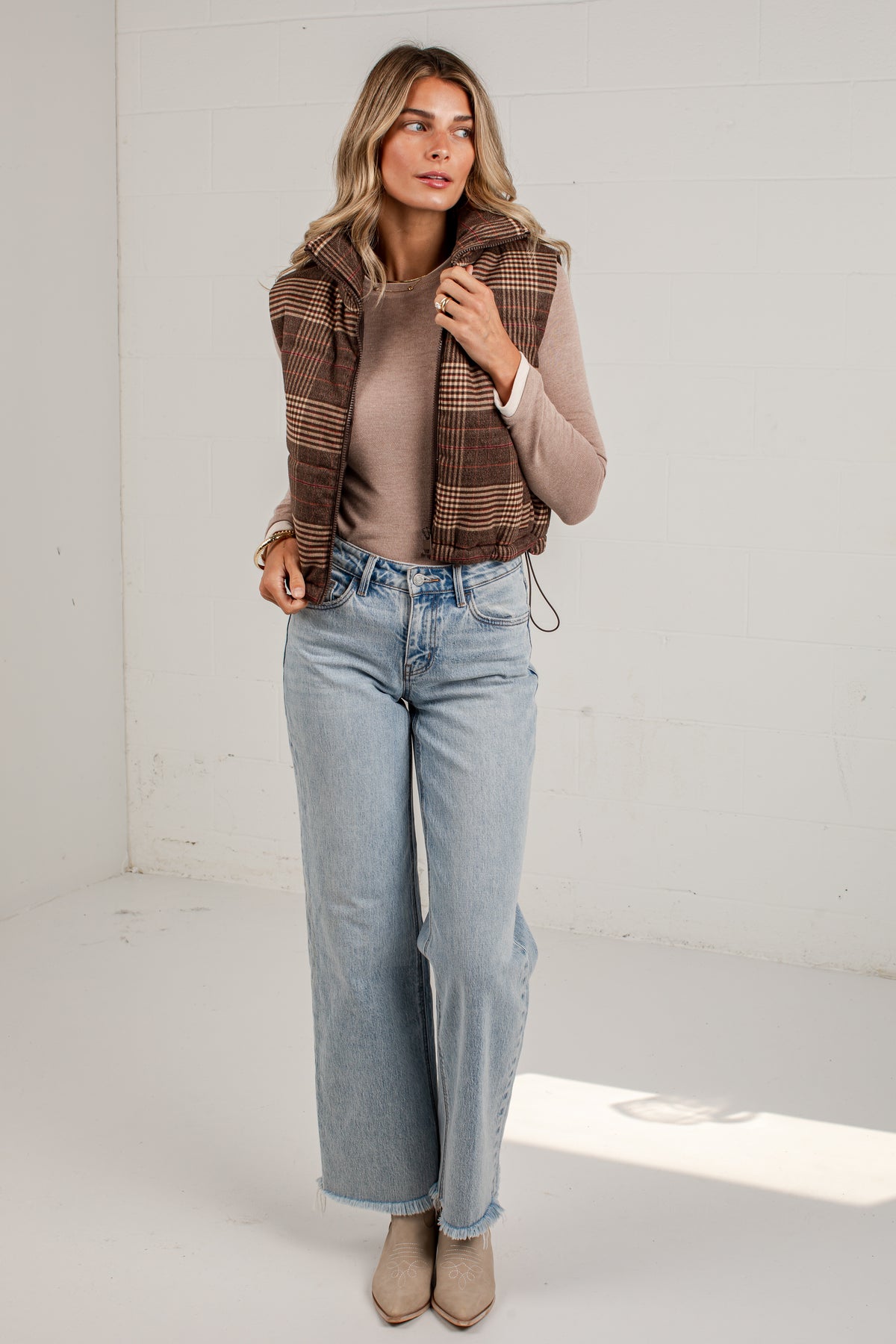 A woman modeling a brown and beige plaid sleeveless vest over a beige top, paired with light-wash wide-leg jeans and beige ankle boots, standing in a bright white studio.